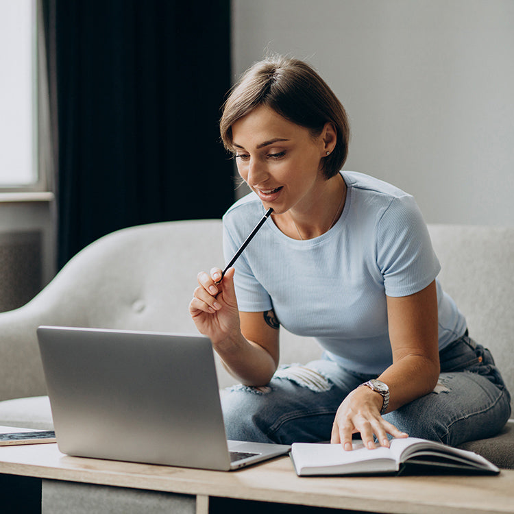 Woman sitting on a couch with a laptop and book, holding a pencil to her chin and appearing to be in deep thought.