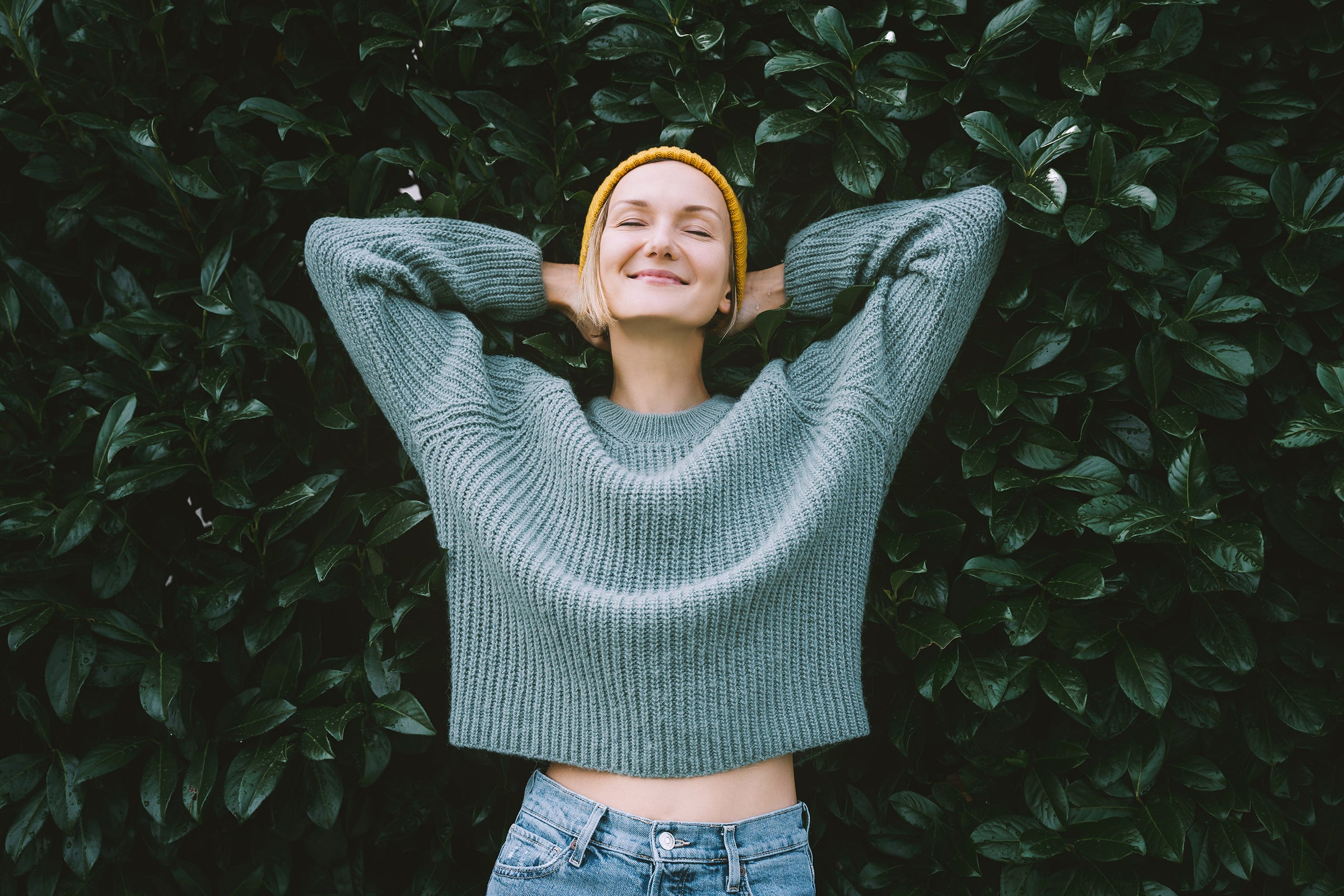 Woman wearing a green sweater and yellow beanie standing against a leafy background.