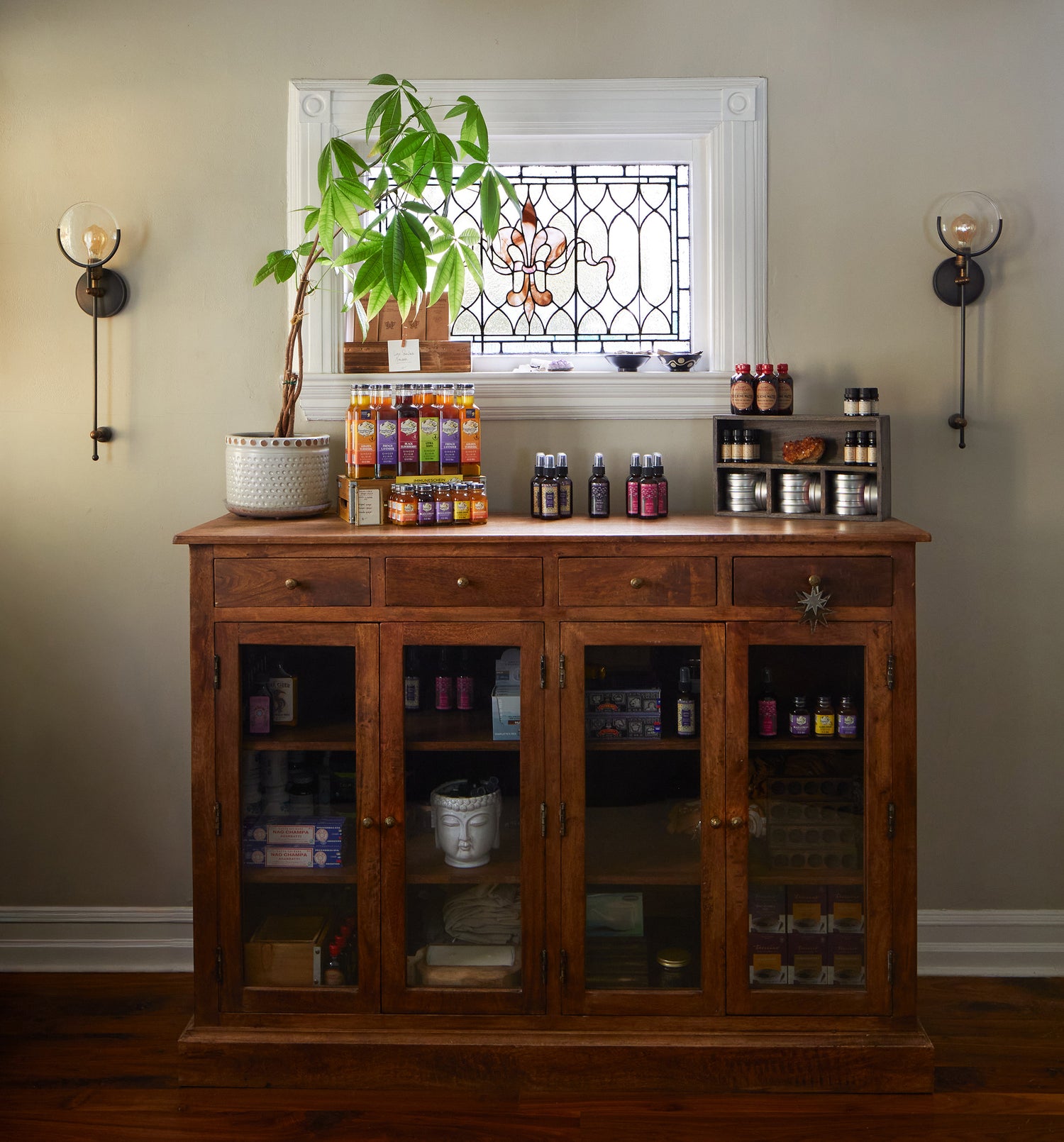 Jennifer Layne reception room merch area, with a stained glass window on the wall, and a wooden cabinet containing various wellness products, mostly in bottles