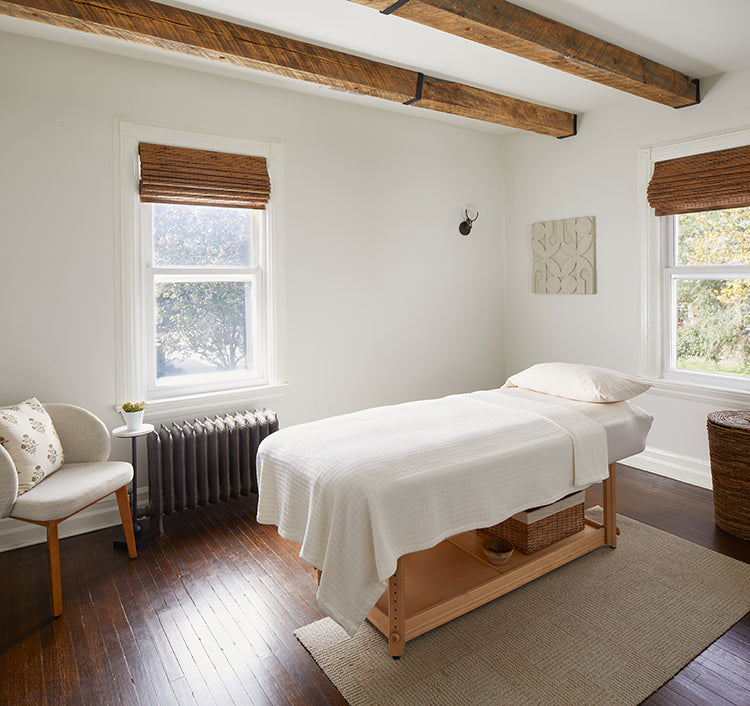 A JWell treatment room with white walls, windows, hardwood floor, white ceiling with wood beams, an acupuncture bed, and an area rug.