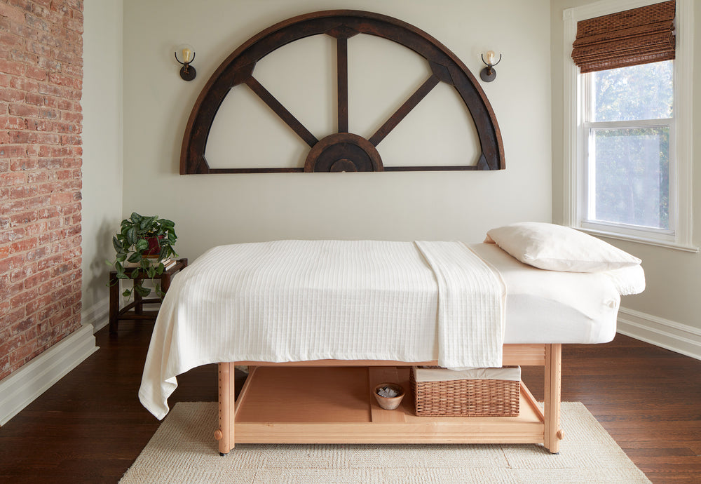 A Jennifer Layne treatment room with acupuncture bed, exposed brick wall, window, hardwood floor, and a large metal half wagon wheel on the wall.