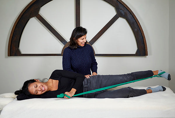 Renuka Pinto guiding a patient who is lying on her side on a physical therapy bed and doing leg exercises with a resistance band.